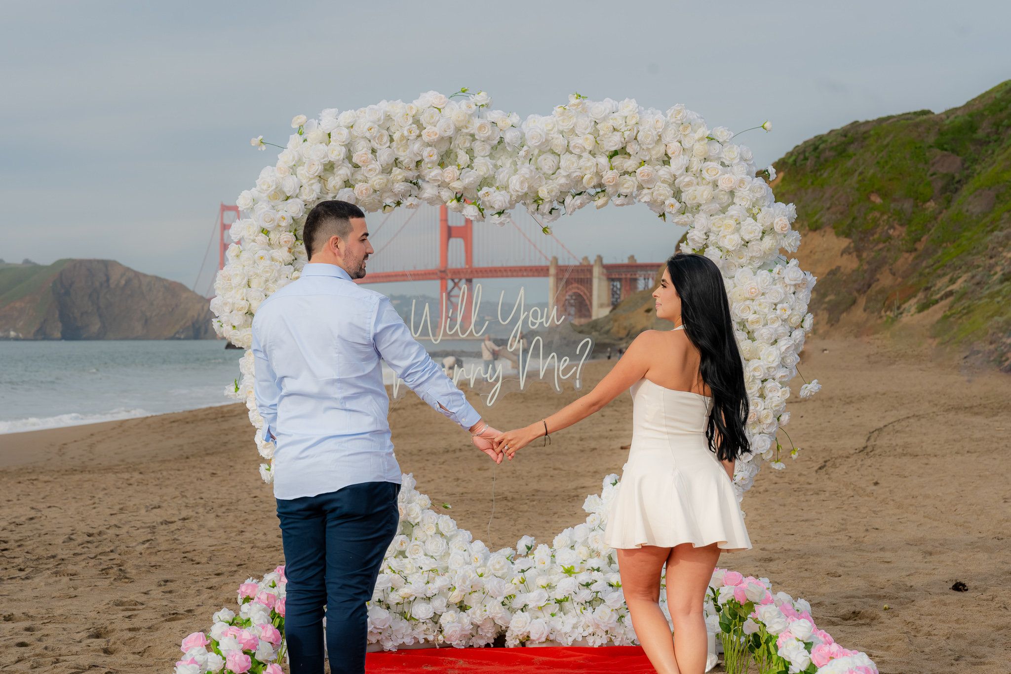 White heart arch proposal with white/pink flower row at Baker Beach with Golden Gate Bridge