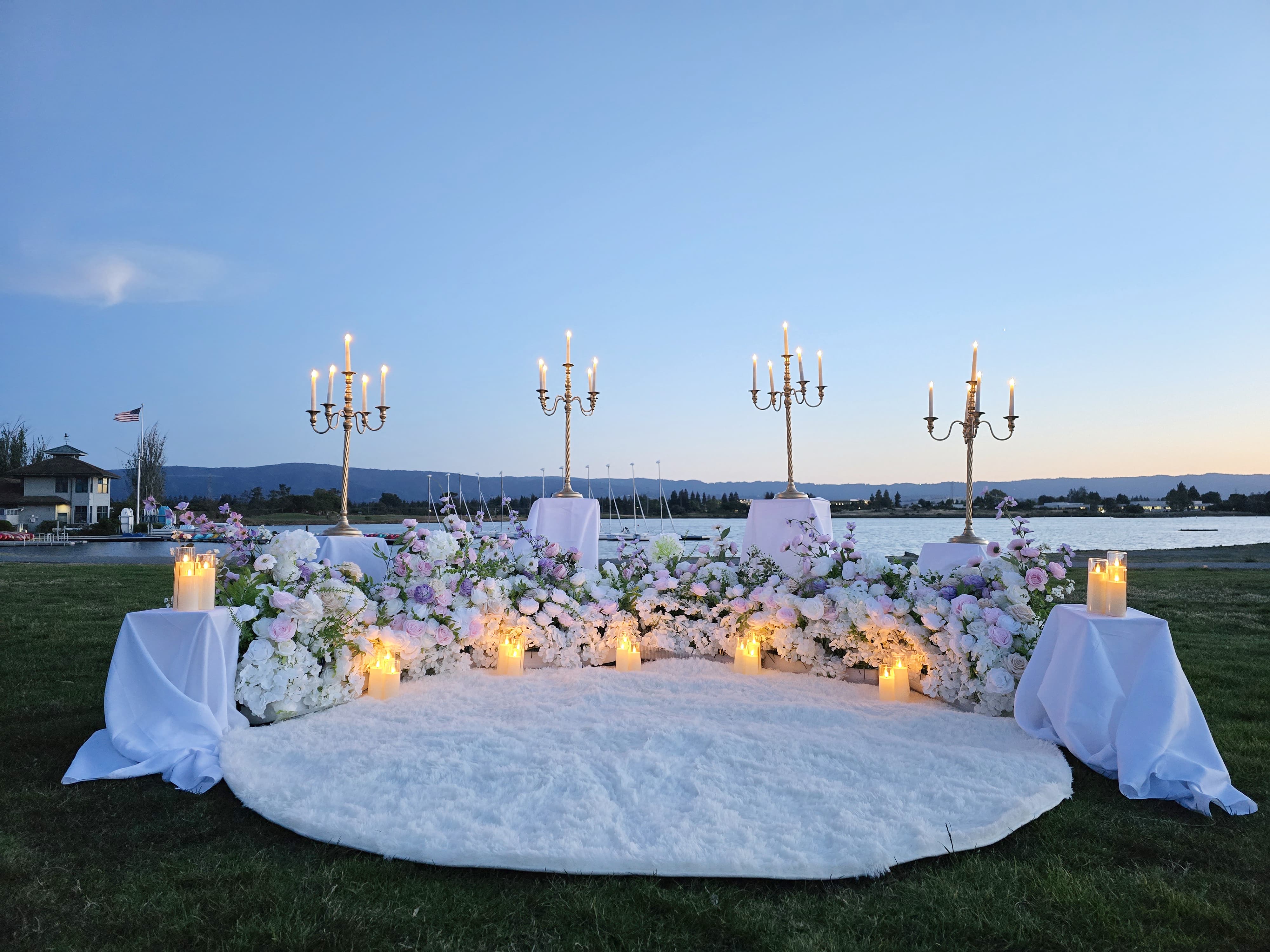 The Enchanted Romance – full view of the proposal setup at dusk with candelabras, florals, and white rug against the bay