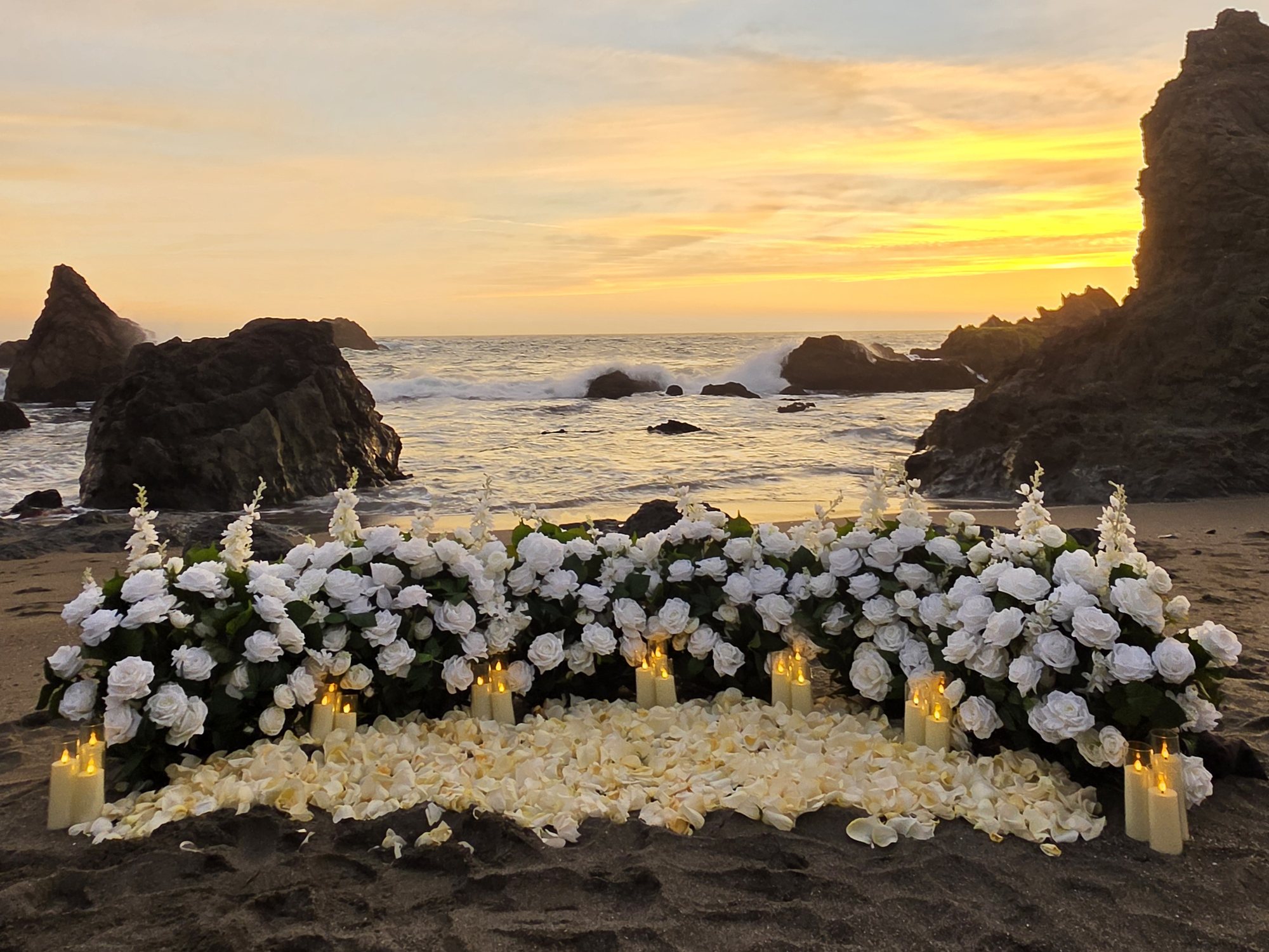 The Intimate Promise proposal setup with white rose grounded florals, rose petals, and candles on the beach at sunset