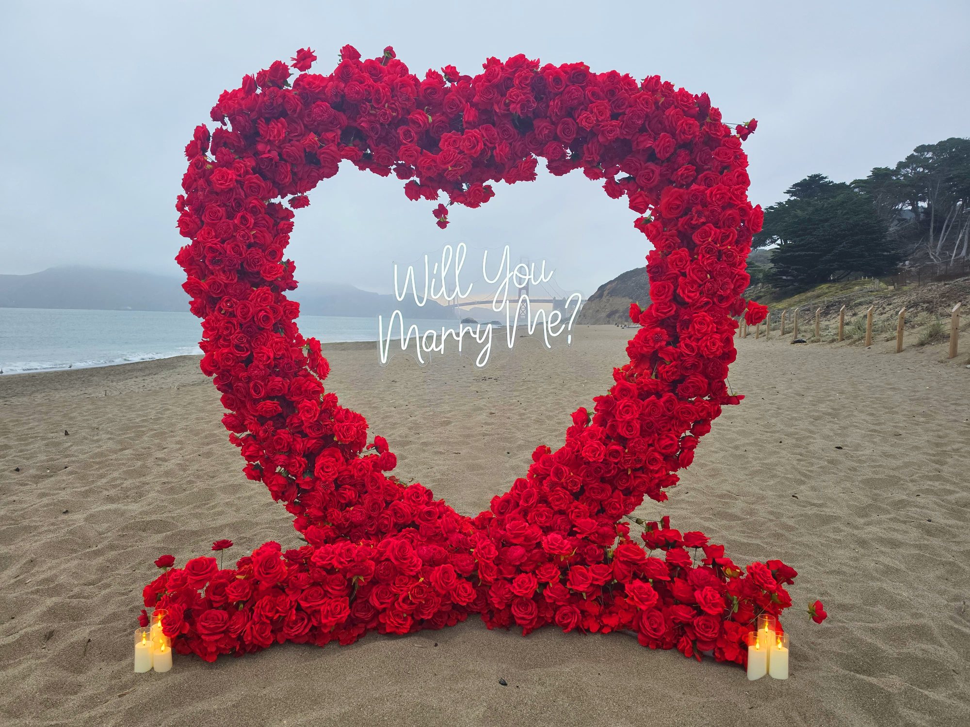 The Little Secret marriage proposal setup – a red rose heart arch with a Will You Marry Me neon sign and pillar candles on the sand at Baker Beach, San Francisco