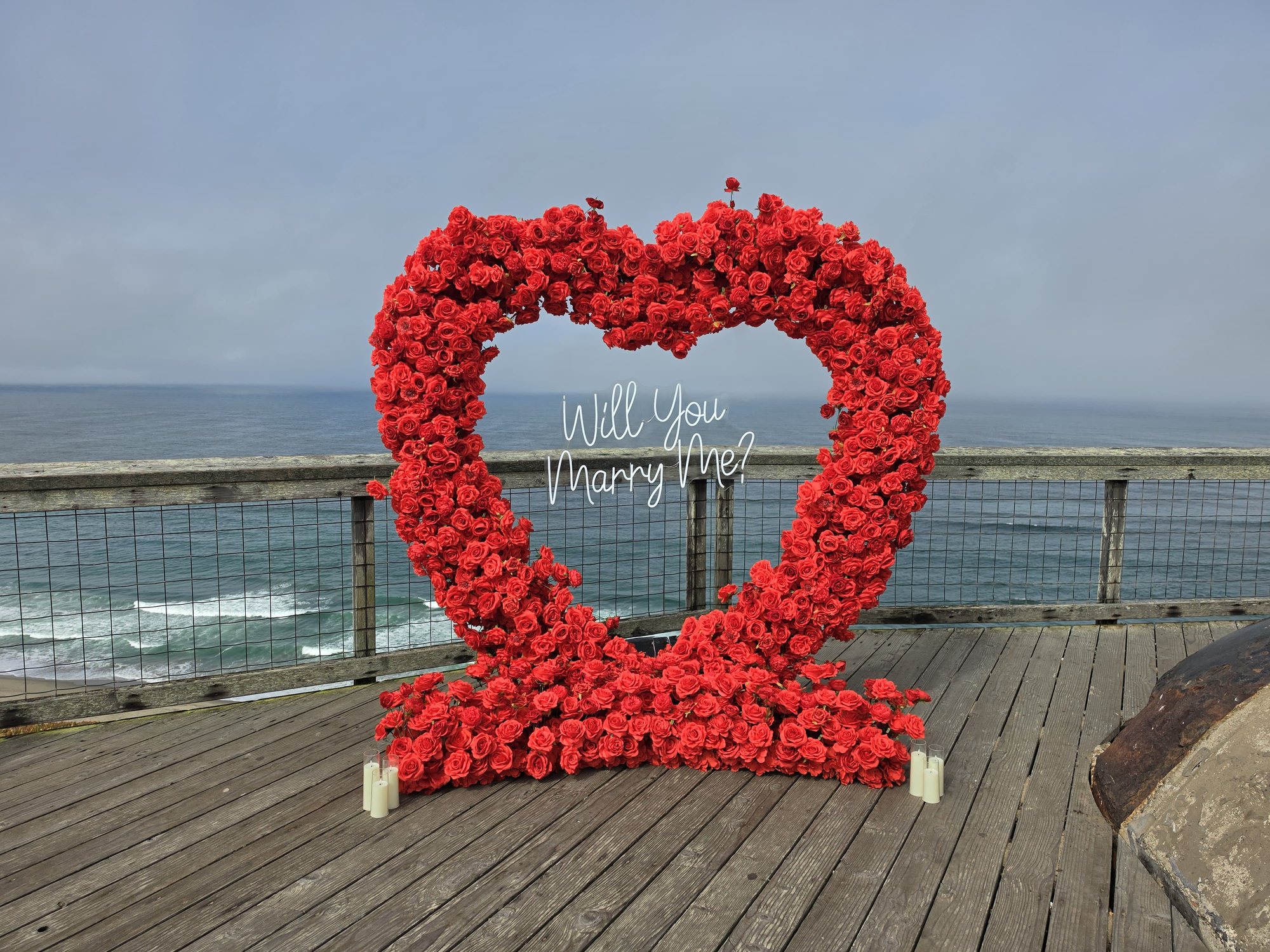 The Little Secret marriage proposal setup – a red rose heart arch with a Will You Marry Me neon sign and pillar candles on the ocean-view deck at Fort Funston, San Francisco
