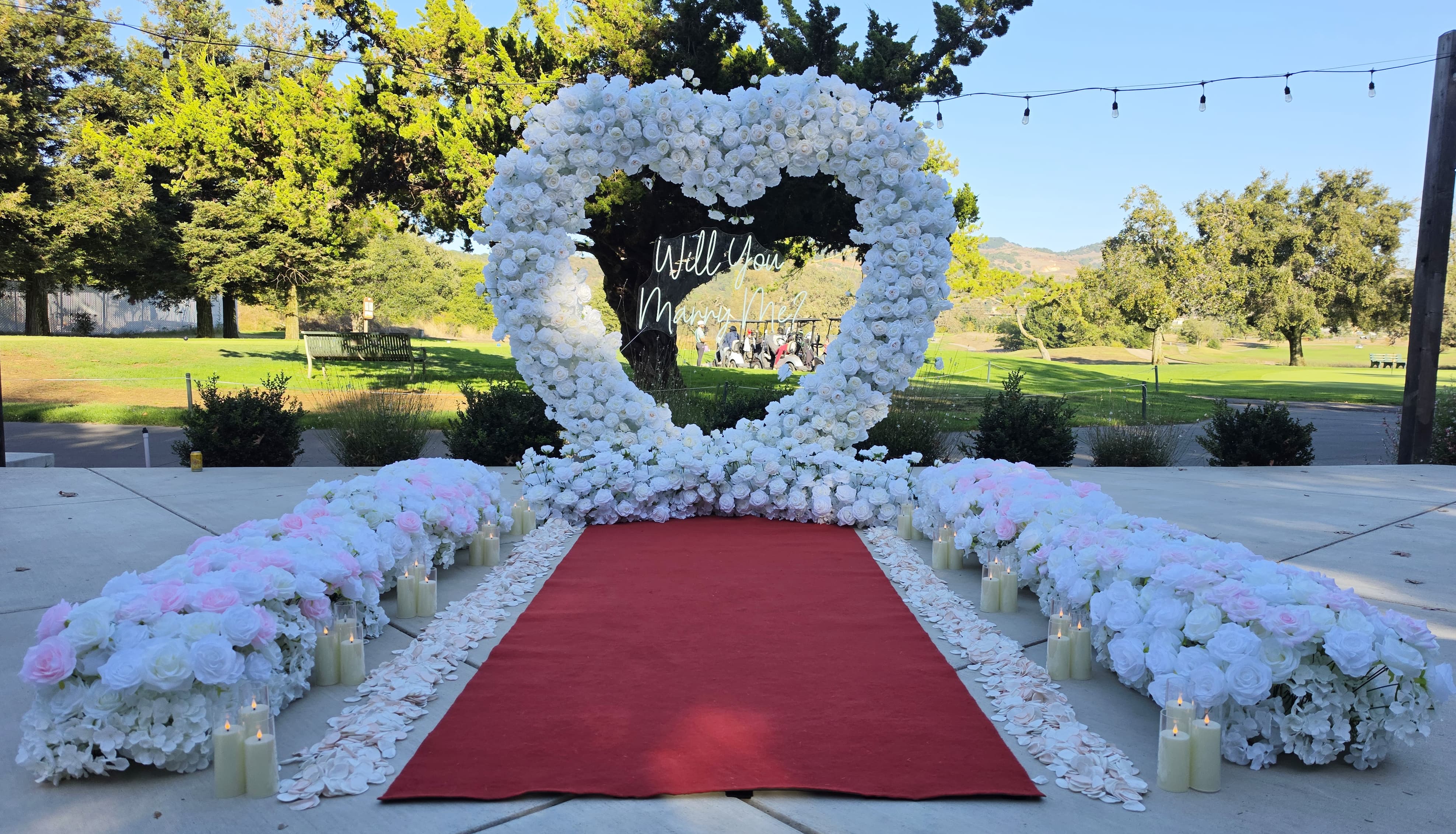 The Masterpiece Package white and pink rose heart arch with "Will You Marry Me?" neon sign, red carpet aisle, and candles at a Napa Valley vineyard