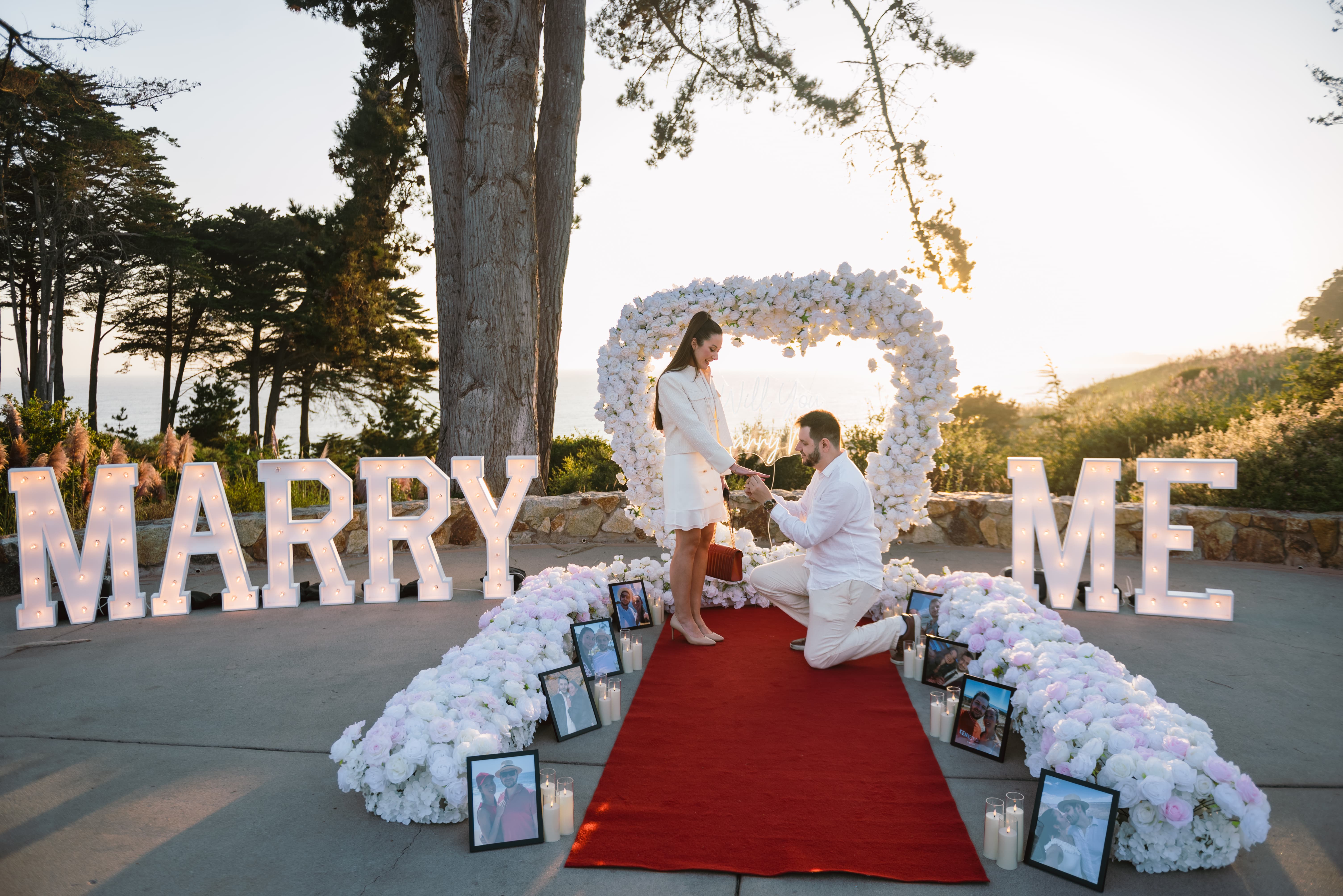 The Grand Beginning Package on the Bluff terrace at Seascape Beach Resort, with a white floral heart arch over the ocean, lit MARRY ME marquee letters, and a red aisle runner lined with white and blush florals and framed memory photos