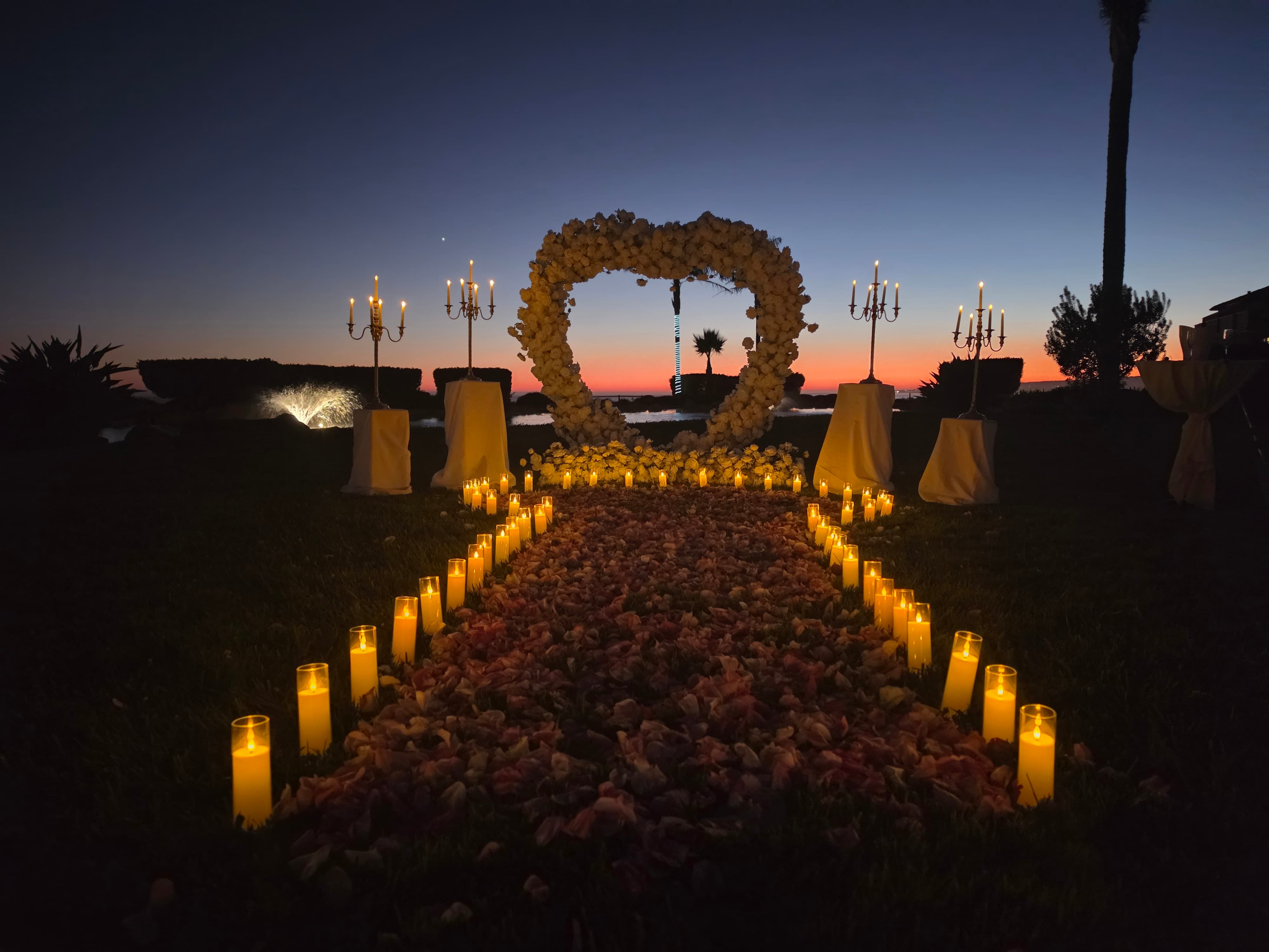 White floral heart arch glowing after sunset on the Island lawn at Seascape Beach Resort, with a candlelit rose petal walkway and gold candelabras