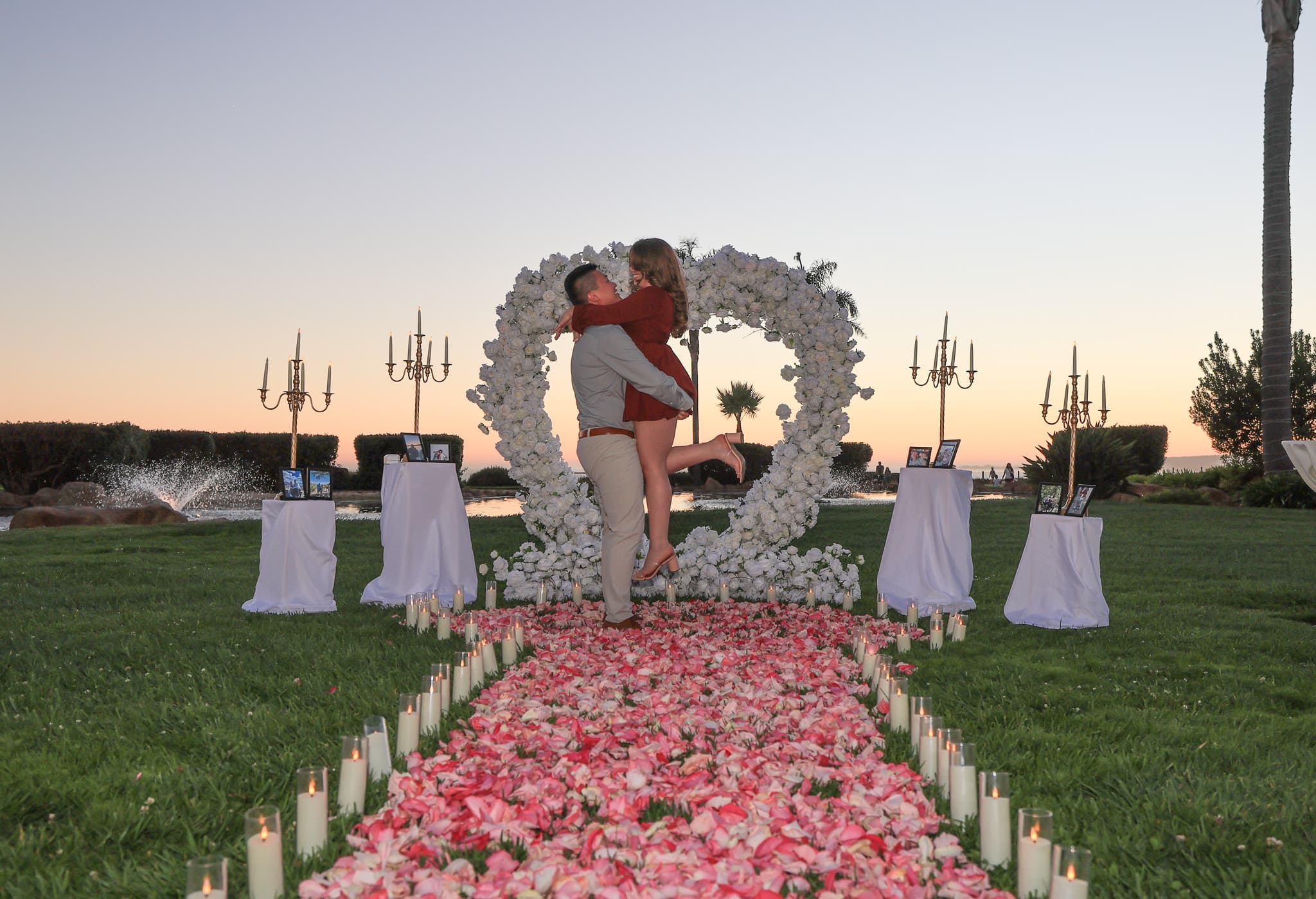 Sunset proposal setup on the Island lawn at Seascape Beach Resort with a heart floral arch, framed photo pedestals, and a petal and candle walkway